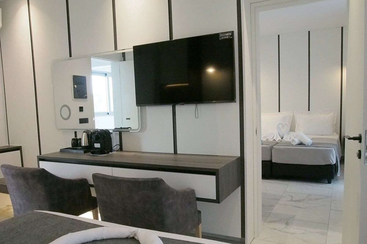 Una habitación de hotel moderna con una televisión de pantalla plana, un escritorio con cafetera, una cama con edredón blanco y un baño visible a través de una puerta abierta.