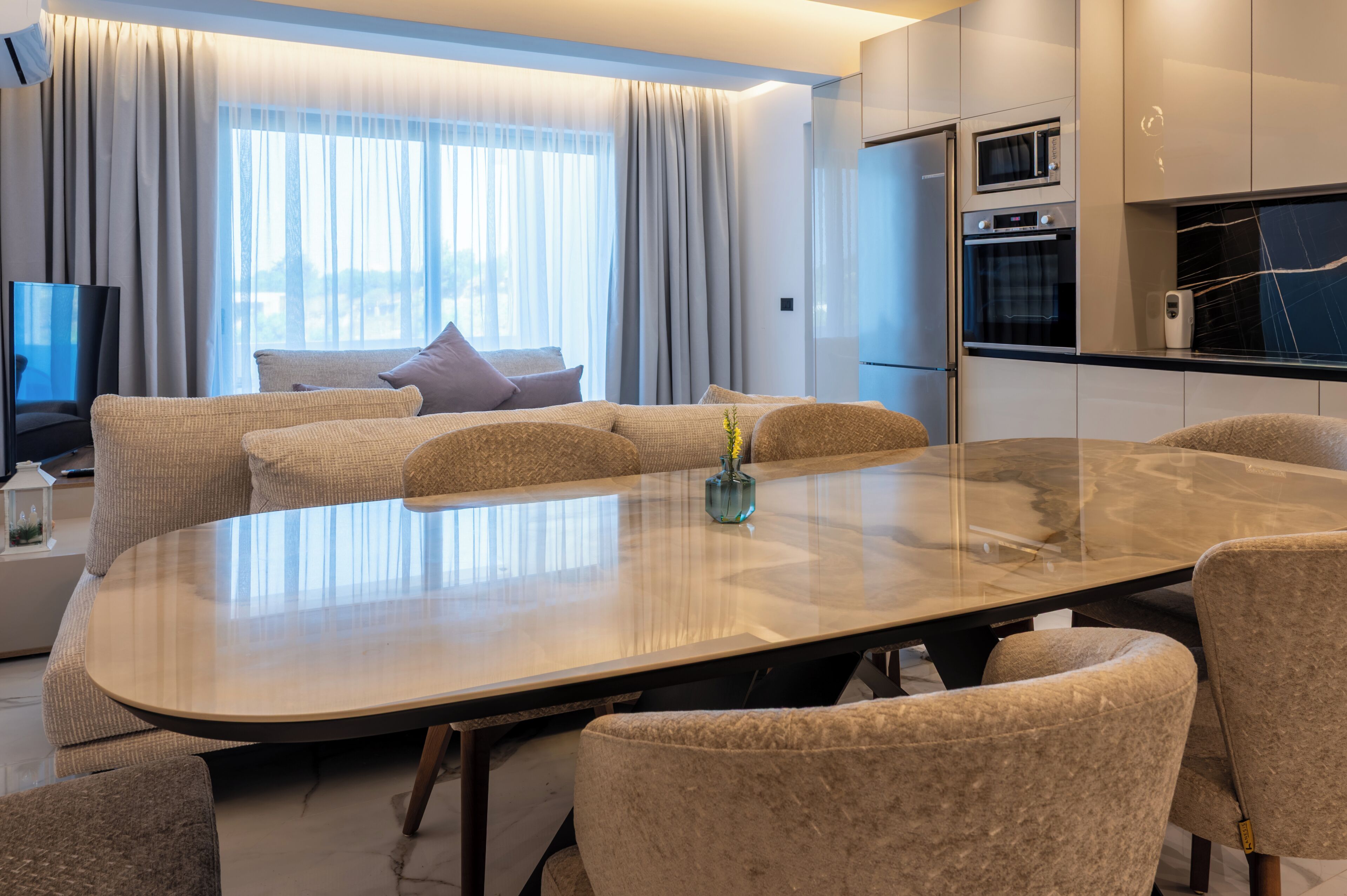 A modern living room with a large glass-top dining table, comfortable seating, a flat-screen TV, and a kitchen area with appliances.