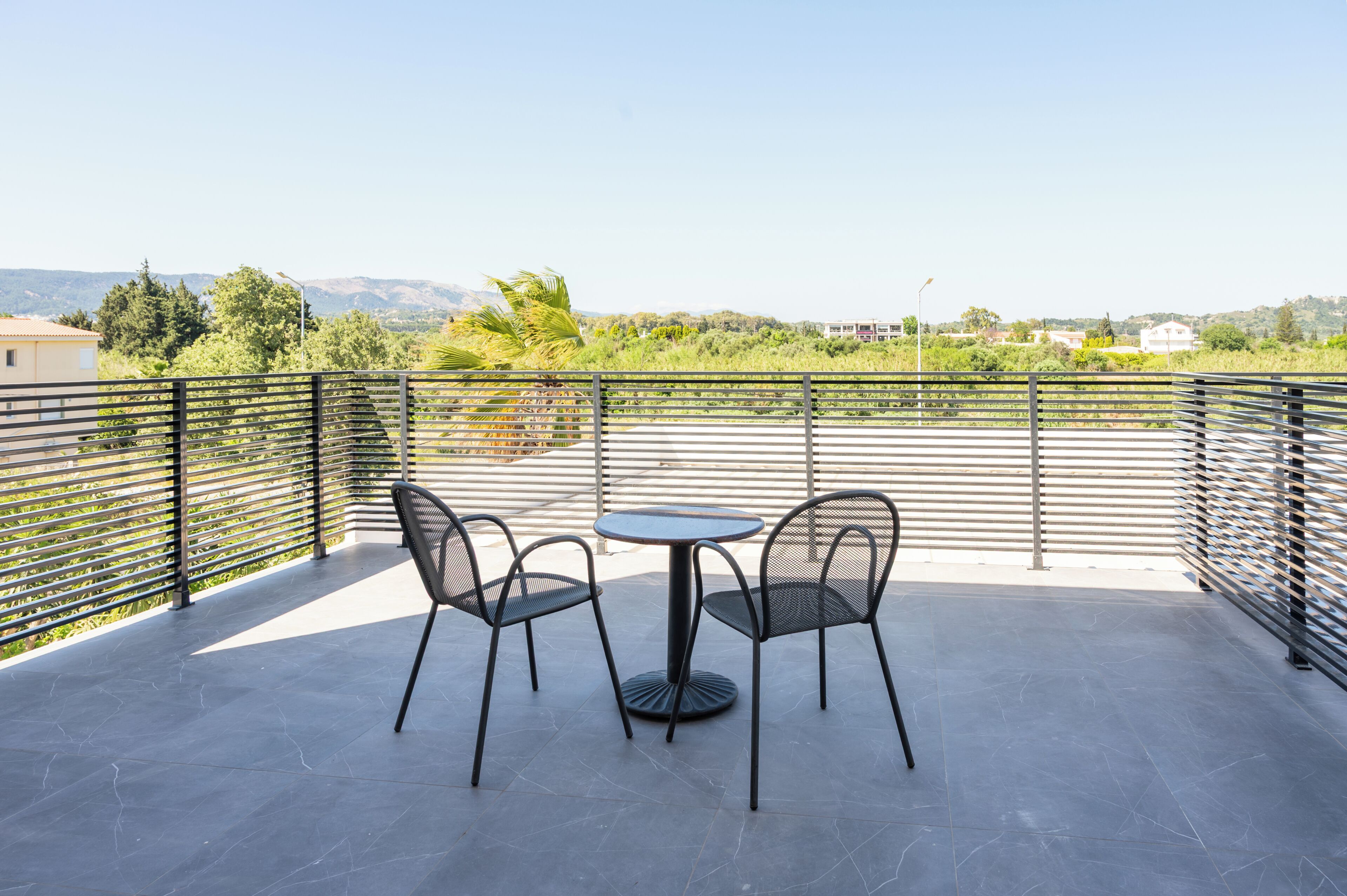 Una terraza con una mesa y sillas, baranda metálica y vistas a zonas verdes y edificios a lo lejos.