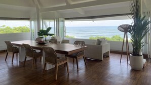 Ein großzügiger Essbereich mit Holztisch und Korbstühlen, große Fenster mit Blick auf das Meer und ein Topfpflanze.