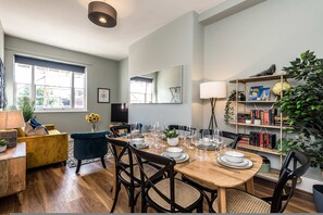 Un comedor con una mesa de madera preparada para cuatro personas, rodeada de sillas negras con asientos de rejilla. A la derecha, una estantería con libros y objetos decorativos.