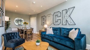 A living room with a blue sofa, a dark armchair, and a wooden coffee table. There is a dining area with a table and chairs, and a wall with the word "ROCK" spelled out in geometric shapes. - Host Stay Catharine Quarters (Liverpool)