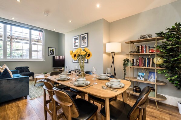 Comedor con mesa de madera dispuesta para seis personas, rodeada de sillas negras. Se observa una estantería con libros y objetos decorativos, y un ventanal que proporciona luz natural.