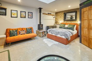 A modern bedroom with a stone accent wall, a bed with a grey fur throw, an orange sofa with patterned cushions, and a small wooden heater.