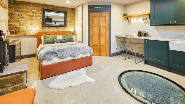 A modern bedroom with a stone wall, a wooden door labeled "RAVENSCAR," a bed with a fur throw, a small kitchen area, and a unique glass floor feature. - Host Stay The Churchside Well (Scarborough)