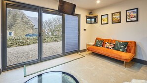 A modern living room with a large sliding glass door leading to a stone building, an orange sofa with decorative pillows, and framed posters on the wall. - Host Stay The Churchside Well (Scarborough)