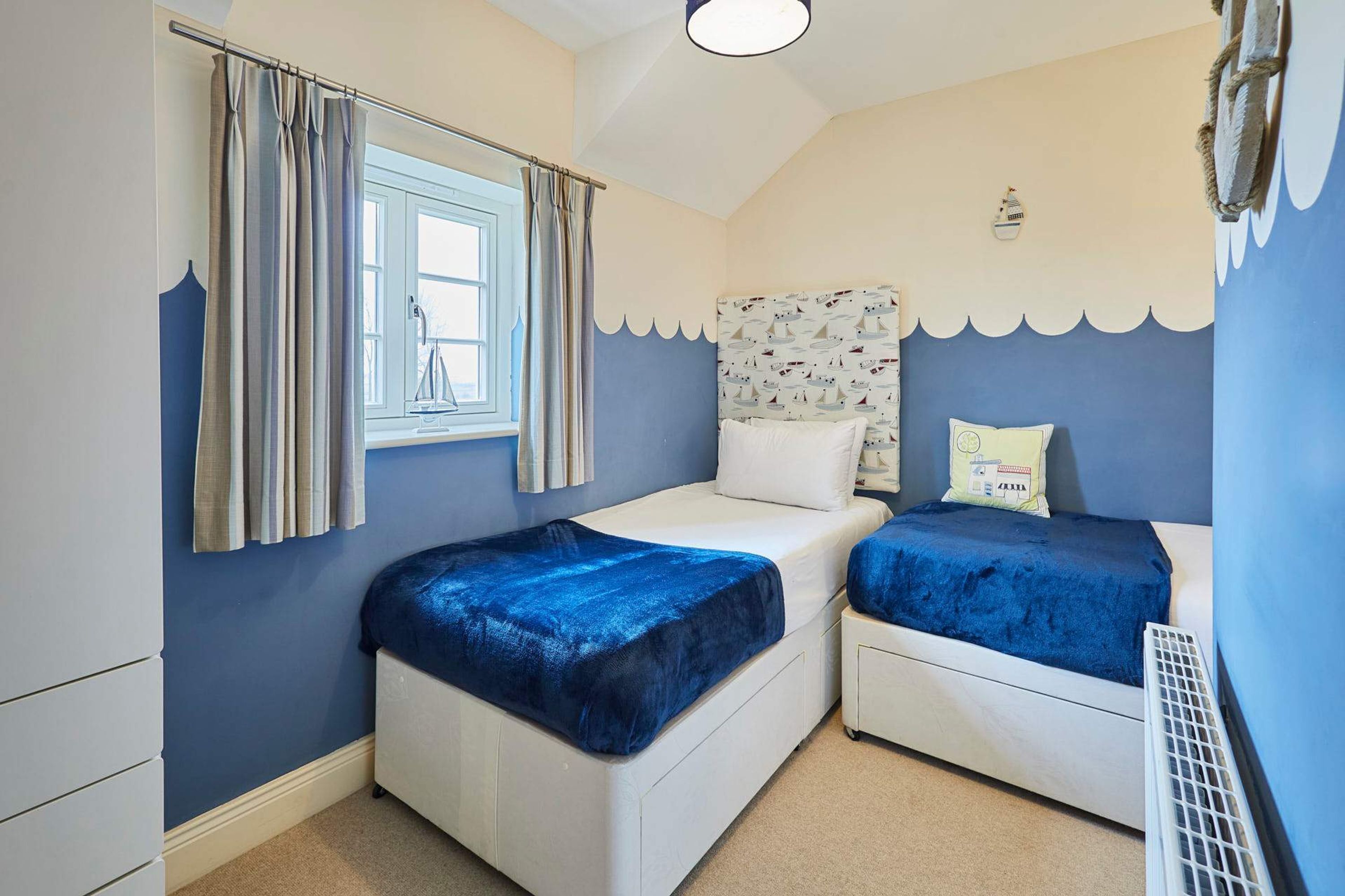 A bedroom with two beds, a window with curtains, a headboard with a nautical pattern, and a radiator.