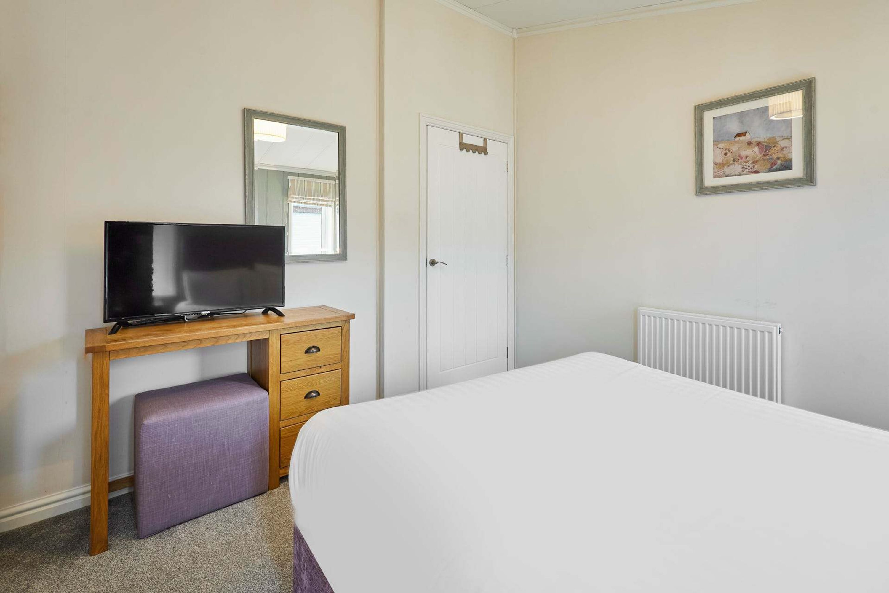 A bedroom with a bed, a wooden desk with a television, a small bench, a door, and a radiator.