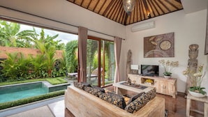 A modern living room with a large window, a pool, and a wooden sofa. - Genie Private Villa by Travellink Bali (Ubud)