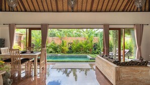 A spacious room with a pool, wooden furniture, and large windows offering a view of lush greenery. - Genie Private Villa by Travellink Bali (Ubud)