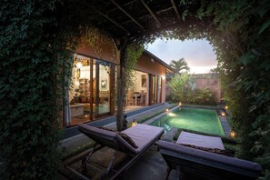 Un espace extérieur paisible avec une piscine et des chaises longues, ainsi qu’un intérieur bien éclairé, visible à travers des portes vitrées.