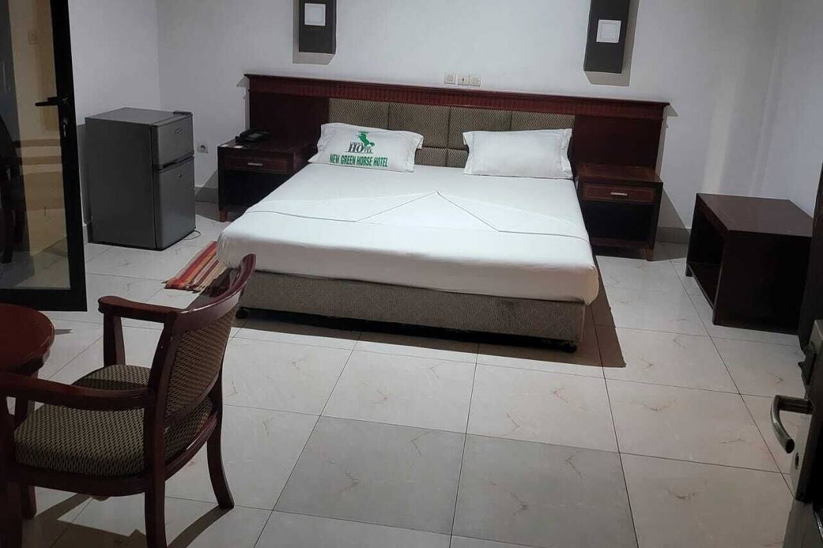 Habitación de hotel con una cama grande, una mesita de noche de madera, una silla, una pequeña mesa y un refrigerador.