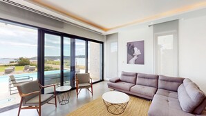 A modern living room with a large window, a grey sofa, two armchairs, a coffee table, and a painting on the wall. - Periyiali Resort - Front Line Villa 08 Chronos (Paralimni)