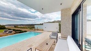 A modern outdoor pool area with a view of the landscape, a seating area with a table, and a large window. - Periyiali Resort - Front Line Villa 08 Chronos (Paralimni)