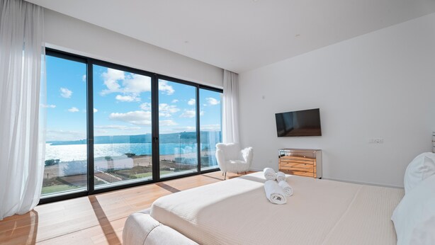 A modern hotel room with a large bed, a flat-screen TV, a wooden dresser, and a view of the sea through floor-to-ceiling windows. - Periyiali Resort - Front Line Villa 08 Chronos (Paralimni)