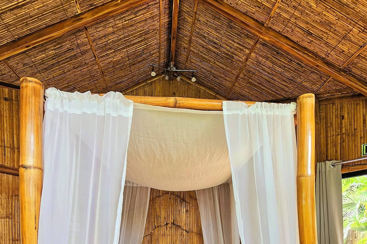 A four-poster bed with white linens and a heart-shaped towel arrangement, set in a room with bamboo walls and a thatched roof.