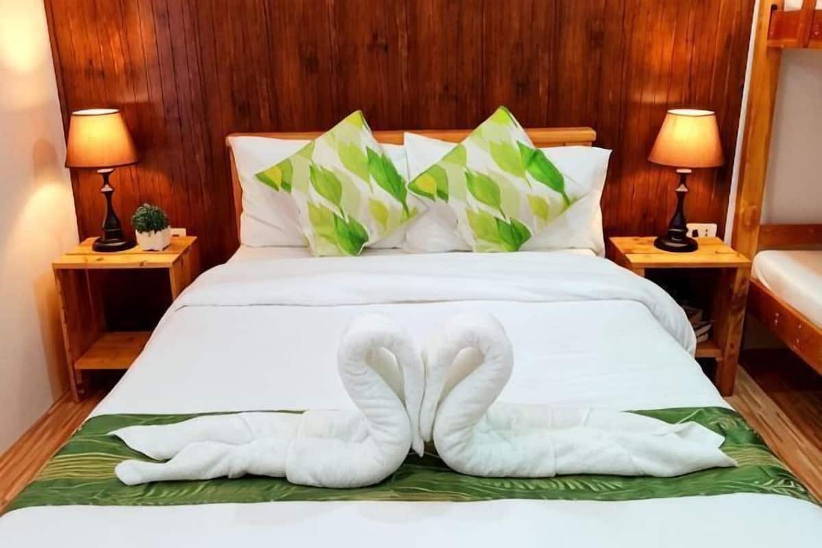 A hotel room with a wooden headboard, two bedside lamps, a bed with white bedding, green leaf-patterned pillows, and a towel arrangement on the bed.