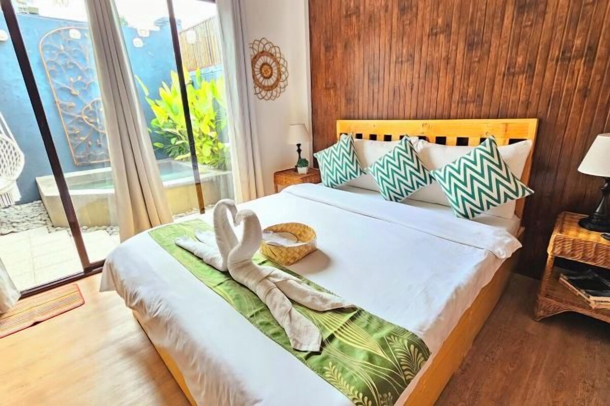 A hotel room with a wooden headboard, a bed with white and green bedding, a pair of towels, a small bedside table, and a view of greenery outside.