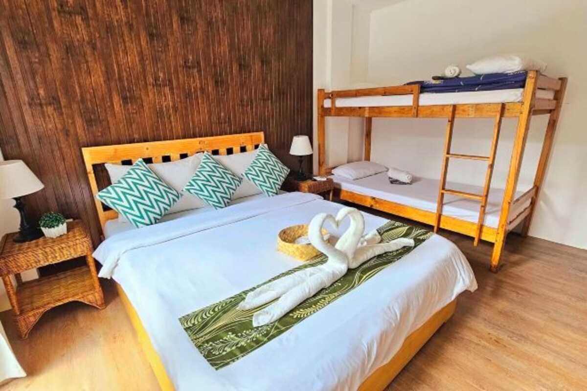 A room with a wooden bunk bed, a single bed with a white bedspread and green leaf-patterned pillow, a small bedside table with a lamp, and a wall with a wooden panel design.