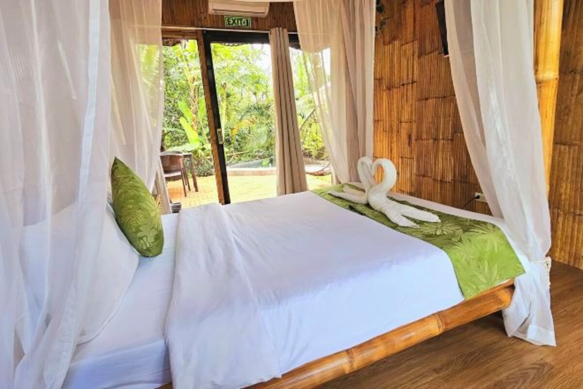 A room with a bed, white curtains, and a view of greenery outside.