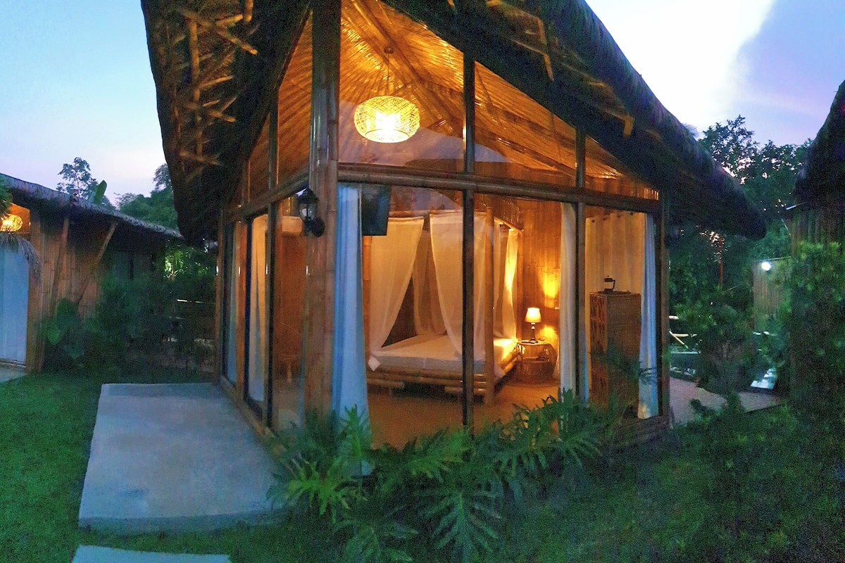 A glass-roofed structure with a bed and bedside lamp, surrounded by greenery.