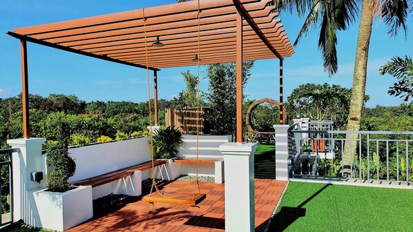 A covered outdoor seating area with a swing, surrounded by greenery and a white fence. - The Farm Shack by SMS Hospitality (Amadeo)