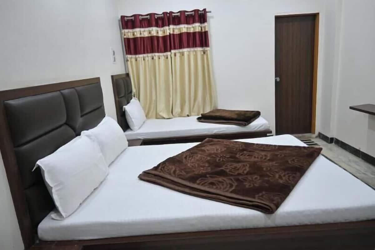 A double bed with a brown blanket, white pillows, and a headboard. There is a nightstand with a lamp, a window with curtains, and a door.