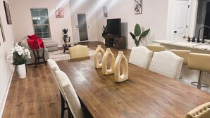 A dining area with a long wooden table, beige upholstered chairs, and a television in the background.