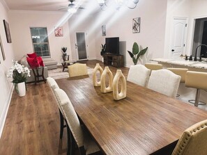 Un coin repas avec une longue table en bois, des chaises recouvertes d’un tissu beige et un téléviseur en arrière-plan.