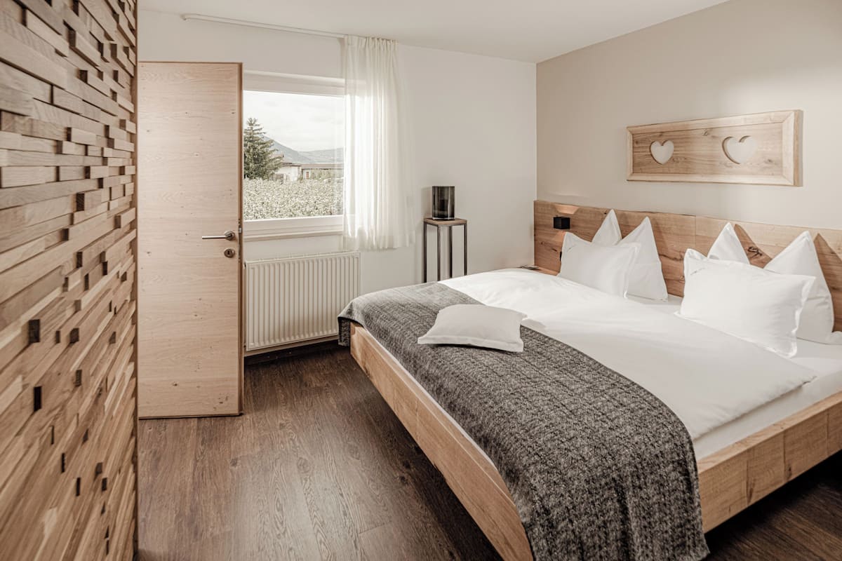 Een slaapkamer met een houten hoofdbord, een groot bed met wit beddengoed, een grijze plaid en een raam met witte gordijnen.