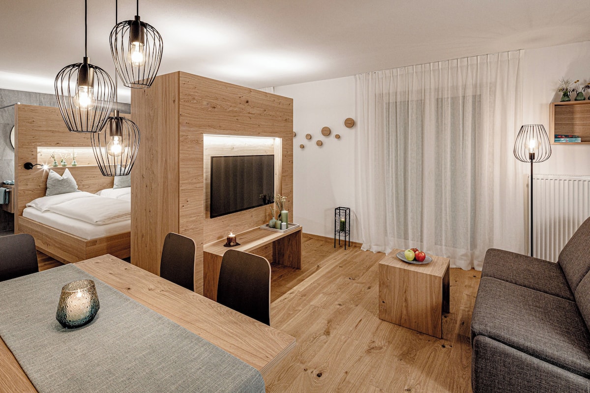 Een moderne hotelkamer met een houten bed, een bureau met een flatscreen televisie, een zithoek met een tafel en stoelen, en een woongedeelte met een bank en een vloerlamp.
