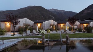 Une rangée de chalets en bois, dotés d’espaces extérieurs aménagés et situés près d’un étang, le tout sur fond de paysage montagneux.