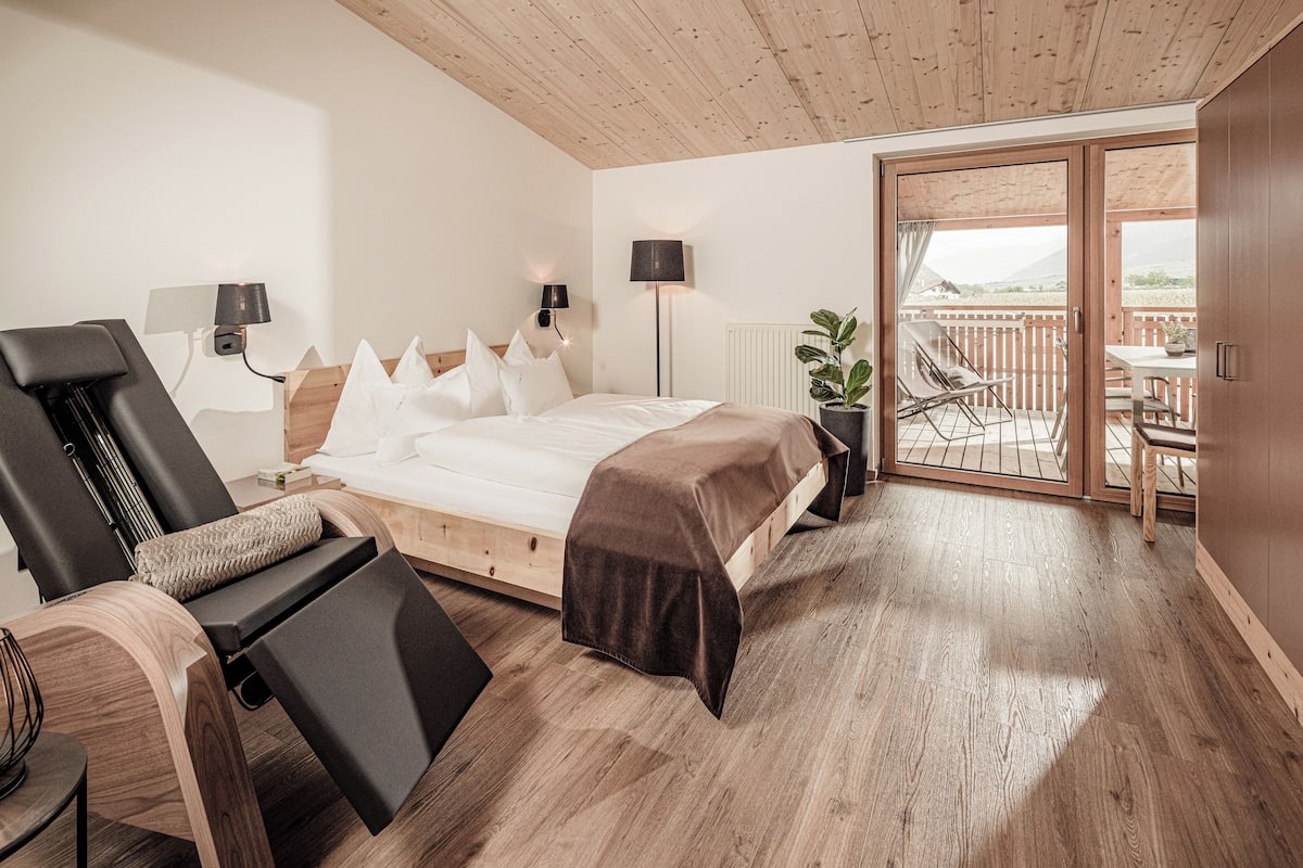 Een slaapkamer met een bed, een fauteuil, een nachtkastje, een lamp en uitzicht op de omgeving door een schuifdeur.