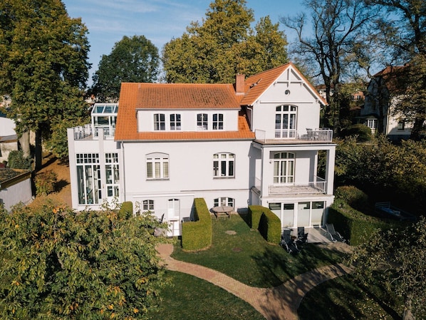 A large, white, two-story house with a red-tiled roof, multiple windows, and a well-maintained lawn.