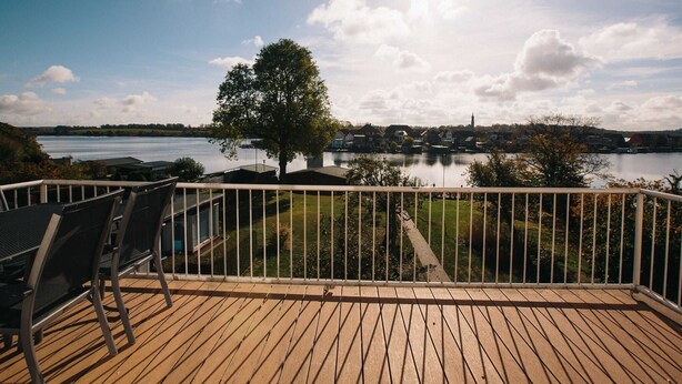 Um terraço com cadeiras de frente para um lago, uma árvore e uma cidade ao longe.