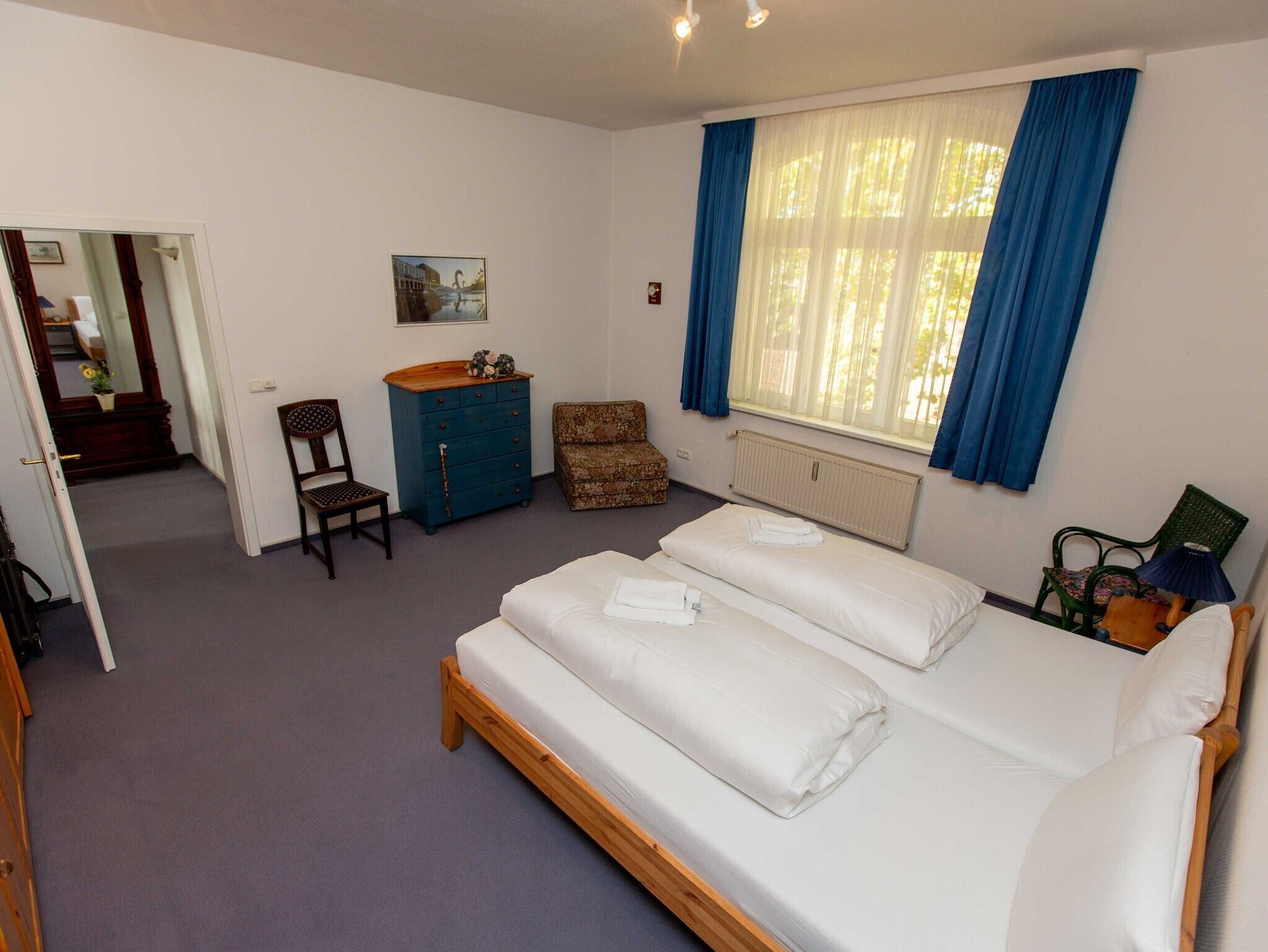 A bedroom with a double bed, a dresser, a chair, a radiator, and a window with curtains.