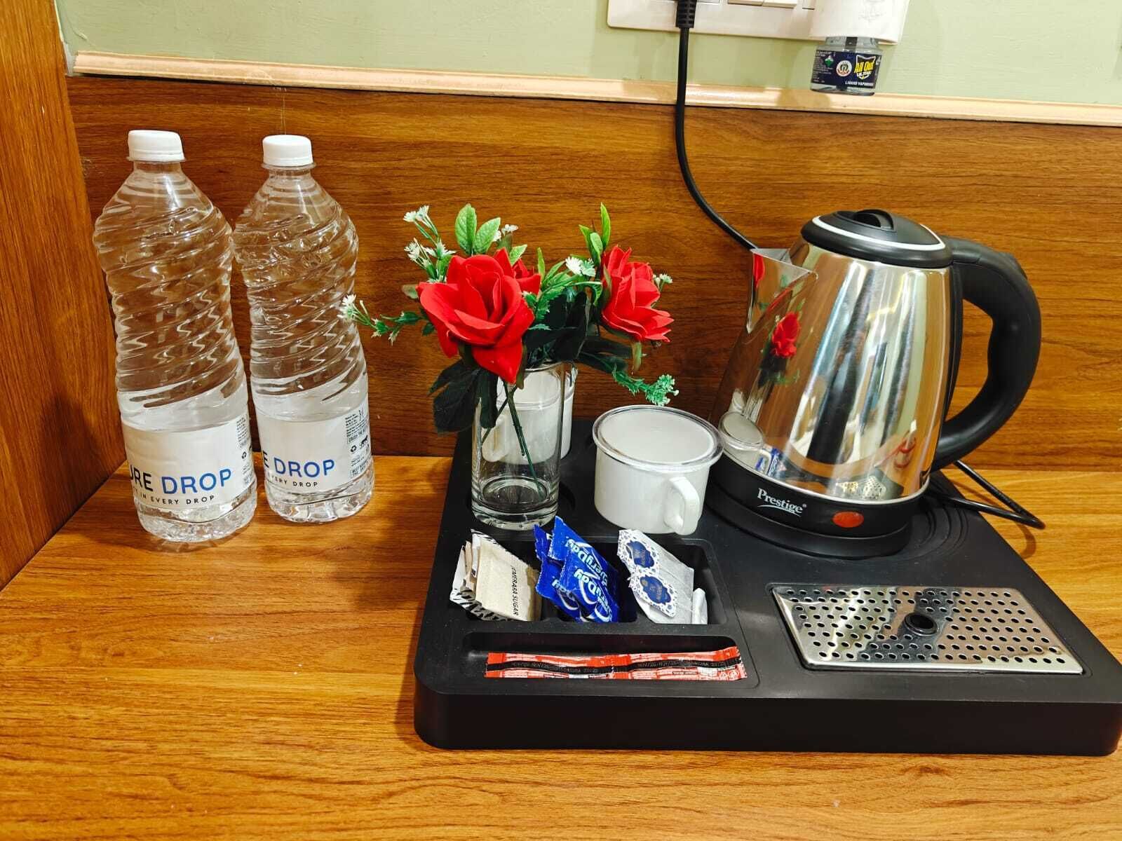 A hotel room's minibar with two water bottles, a vase with artificial flowers, a coffee maker, a cup, and some snacks.