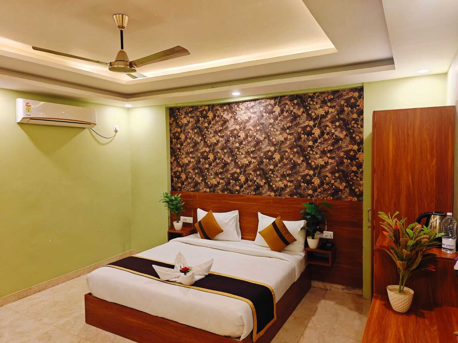 A hotel room with a bed, a ceiling fan, a floral wallpaper, a wooden headboard, and a nightstand with a plant.