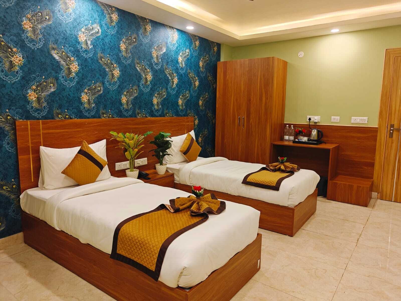 A hotel room with two beds, a wooden wardrobe, a small desk with a kettle, and a decorative wall.