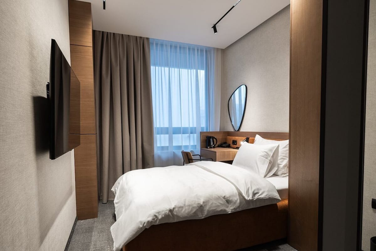 Une chambre d’hôtel moderne dotée d’un grand lit, d’un téléviseur à écran plat fixé au mur, d’une table de chevet avec une lampe et d’une fenêtre ornée de rideaux légers.