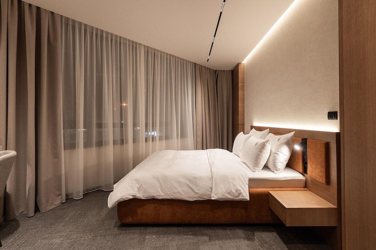 Une chambre d’hôtel moderne avec un grand lit, une tête de lit en bois et une vue dégagée à travers des rideaux légers.