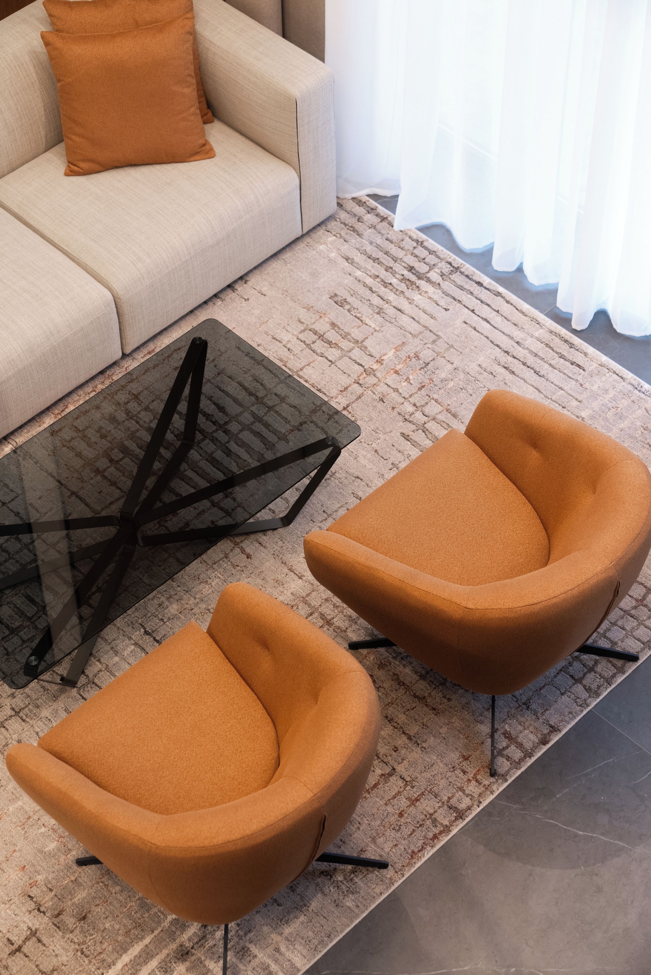 Una sala de estar moderna con un sofá color crema, dos sillones naranjas y una mesa de centro de vidrio.