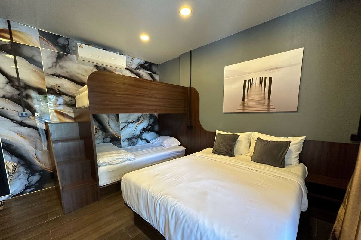 Una habitación de hotel con una cama grande, una cama más pequeña y una litera. Hay un cuadro en la pared y una lámpara en el techo.