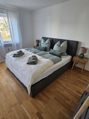 A modern bedroom with a large bed, wooden flooring, and a nightstand with a lamp.