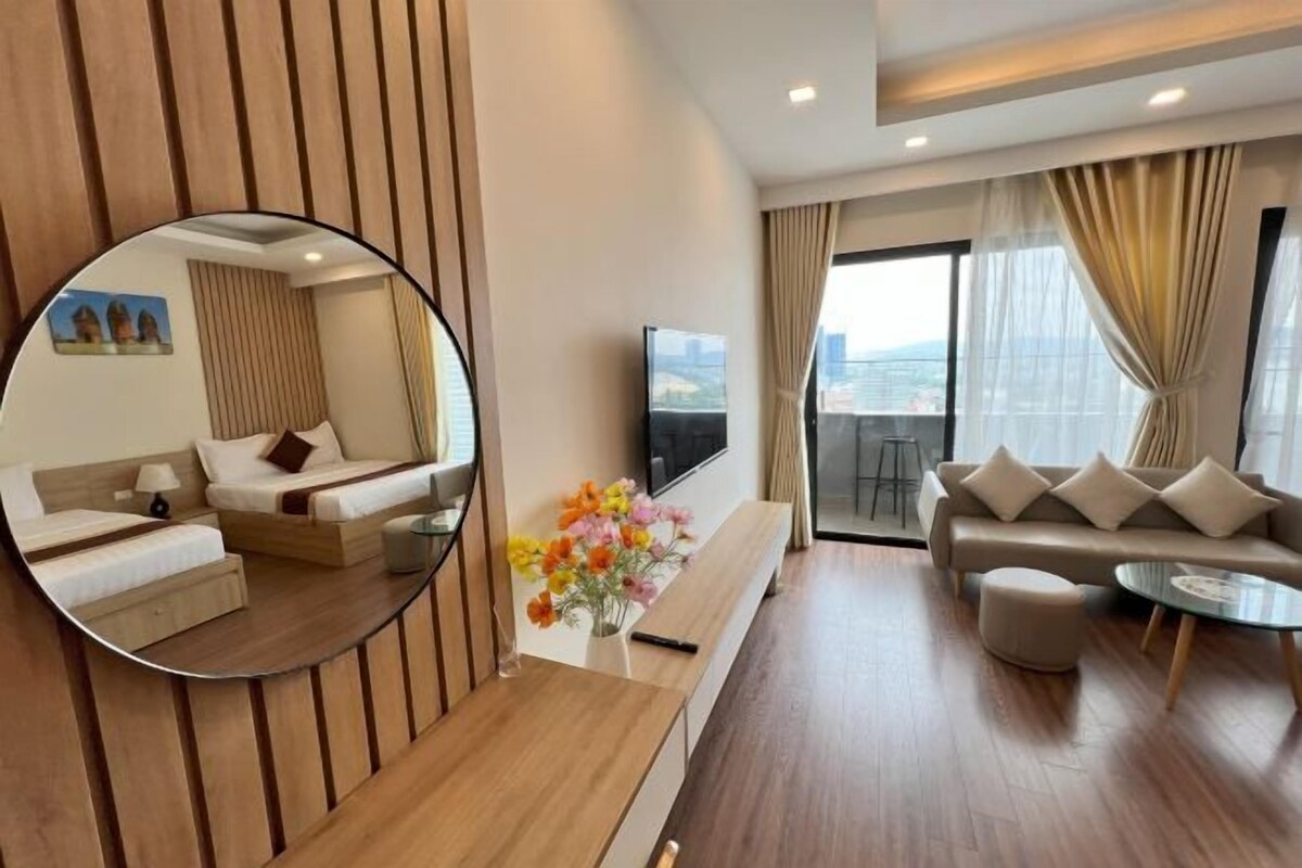 A modern hotel room with a wooden accent wall, a round mirror, a sofa set, a coffee table, a TV, and a balcony with a view.