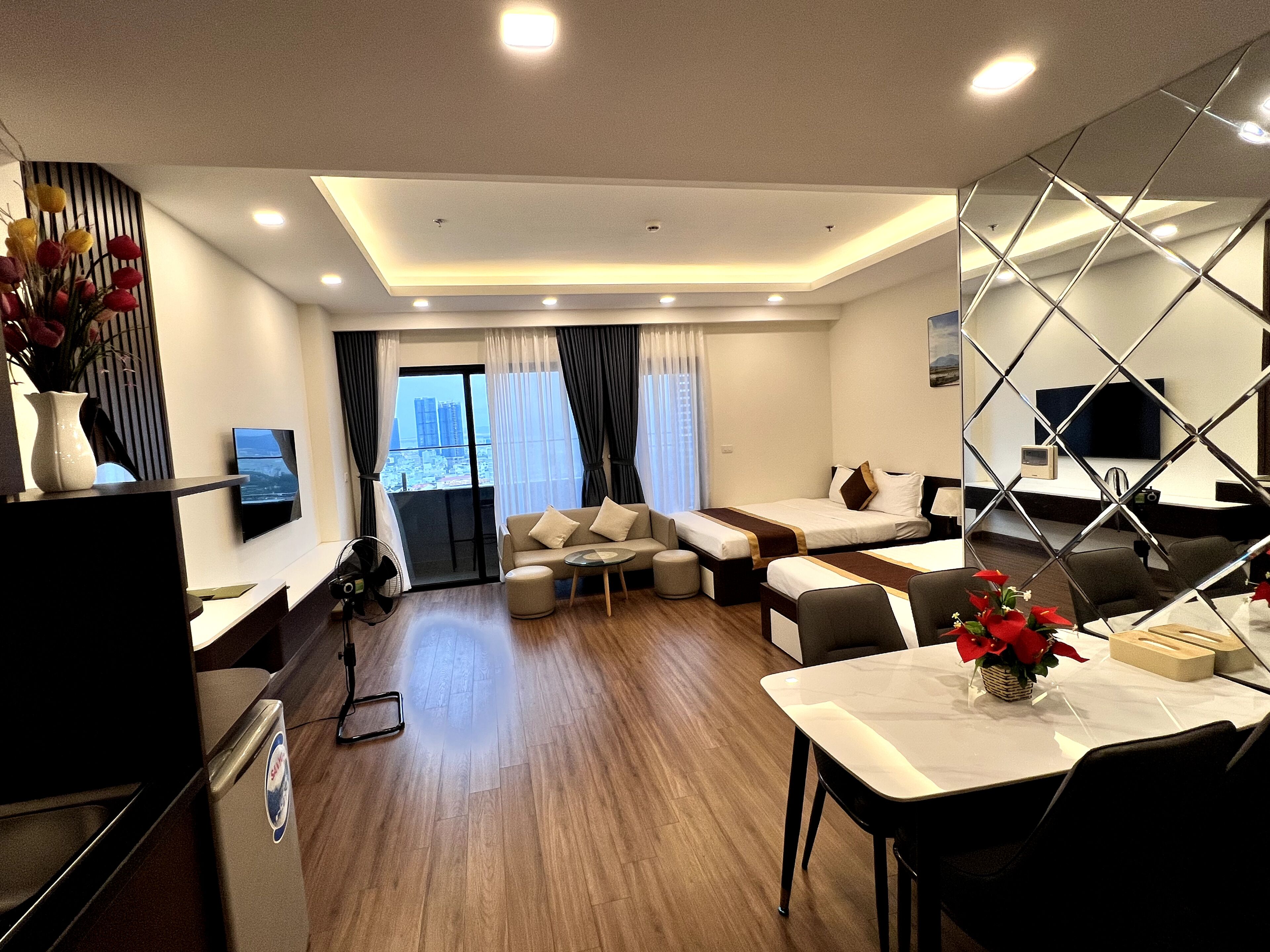 A modern hotel room with a sofa set, a dining table, a TV mounted on the wall, and a view of the city through the window.