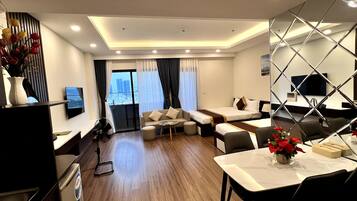 A modern hotel room with a sofa set, a dining table, a TV mounted on the wall, and a view of the city through the window.