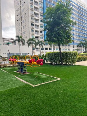 A playground with swings and a tree in a grassy area. - Cozy 1BR at Sea Residences Near MOA - Wi-Fi (Pasay)