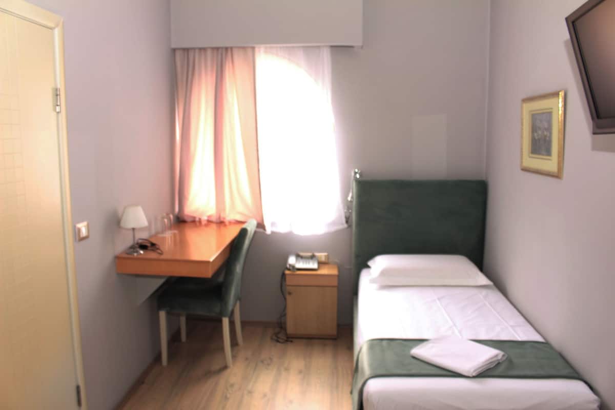 A small, single-bed room with a desk and chair, a bedside table with a lamp, and a window with curtains.