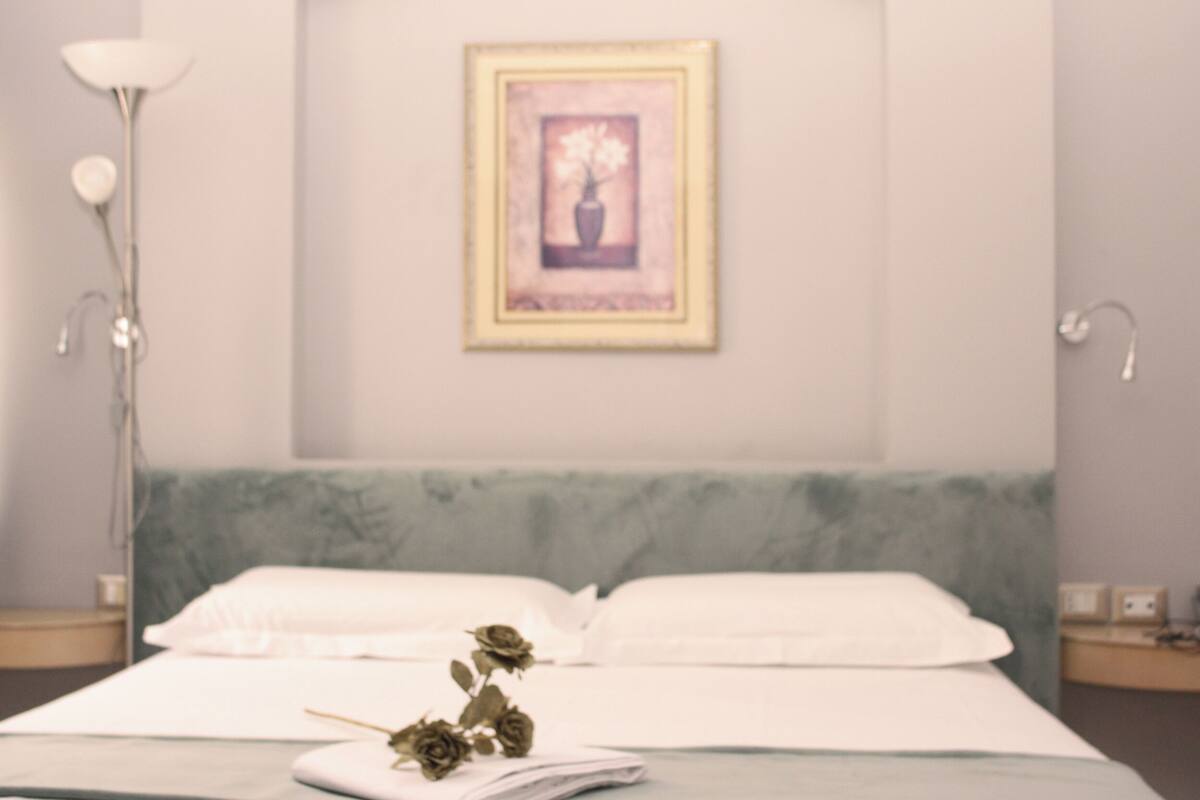 A neatly made bed with white linens, a framed artwork above it, and a bedside lamp on the left.
